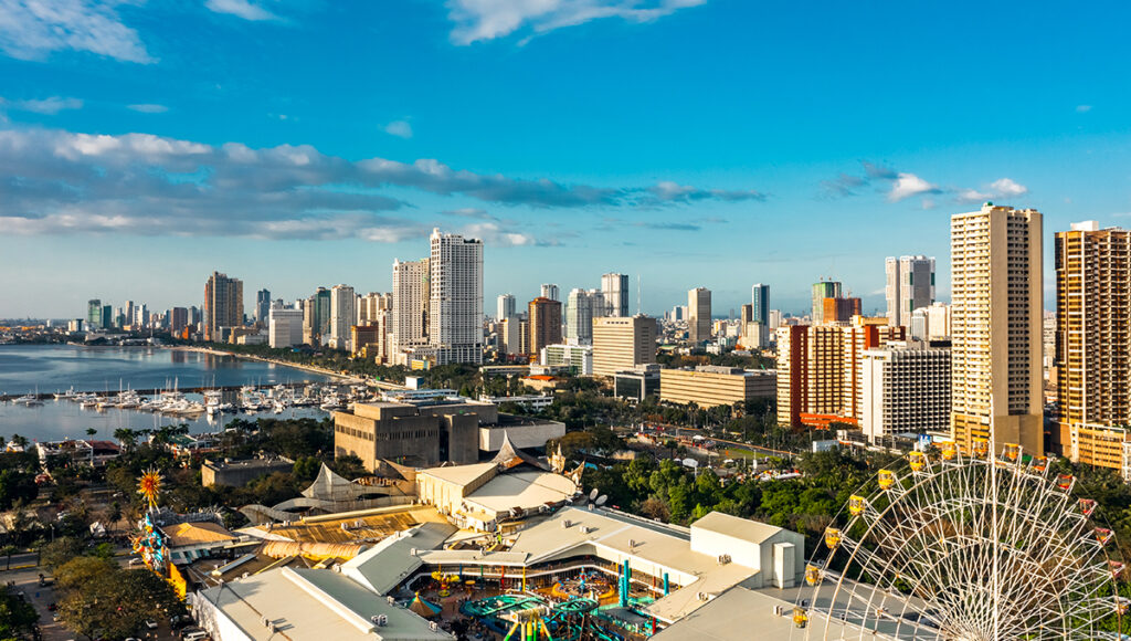 A high-angle, panoramic view of the Manila skyline along the bay, featuring numerous high-rise buildings, a marina with docked boats, and a large amusement park area with a Ferris wheel in the foreground.