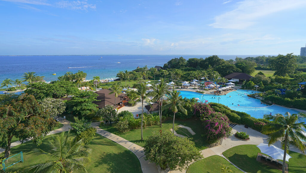 Aerial view of a luxurious, tropical beachfront resort on Mactan Island, Cebu, Philippines, featuring a large swimming pool, lush gardens, and palm trees. This location is a prime example of where to stay in Cebu for a relaxing island vacation.