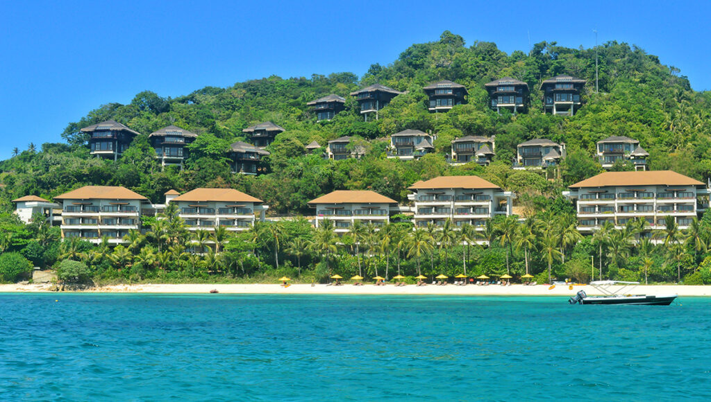 A wide view of the luxurious Shangri-La Boracay resort built into a lush green hillside overlooking a pristine beach. The resort is composed of larger multi-story buildings closer to the beach and smaller, darker villas tiered higher up the forested slope. Turquoise water is visible in the foreground, with a small boat anchored near the shore.