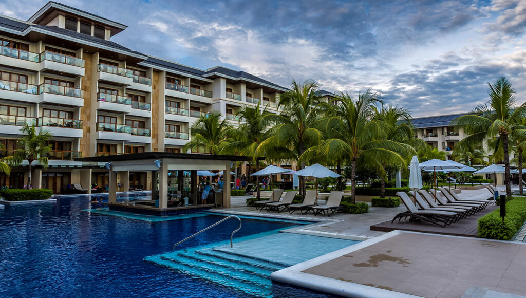 A view of the main swimming pool area at the Henann Resort Alona Beach in Bohol, Philippines. The large blue pool features steps leading to a shallow section and a built-in swim-up bar area. In the background are the modern, multi-story resort buildings and rows of sun loungers and palm trees under a dramatic, cloudy sky.