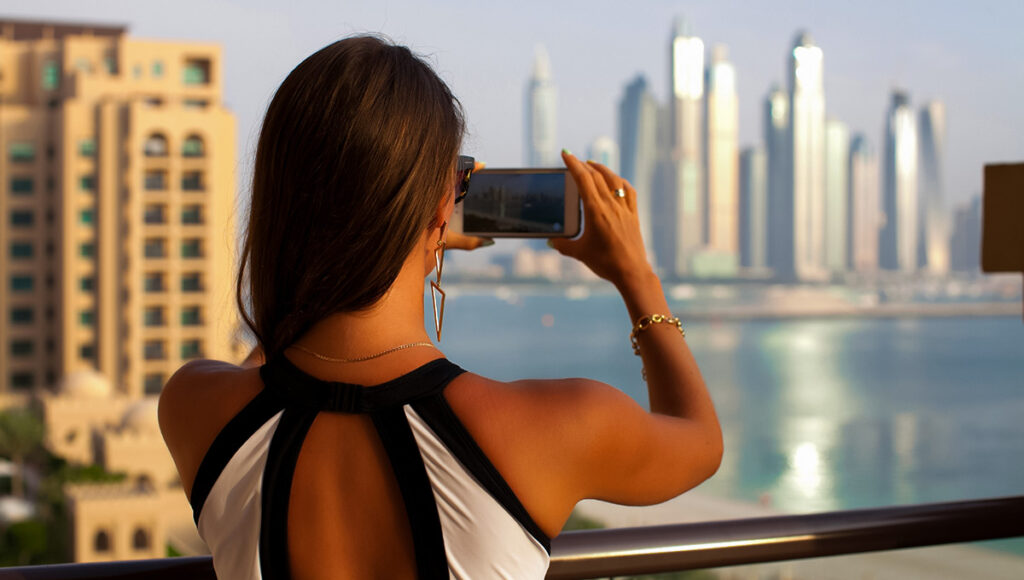A woman, seen from behind, takes a photo with her smartphone of a modern city skyline across a body of water from a high balcony, suggesting a scenic view similar to those found at some all-inclusive resorts Philippines.