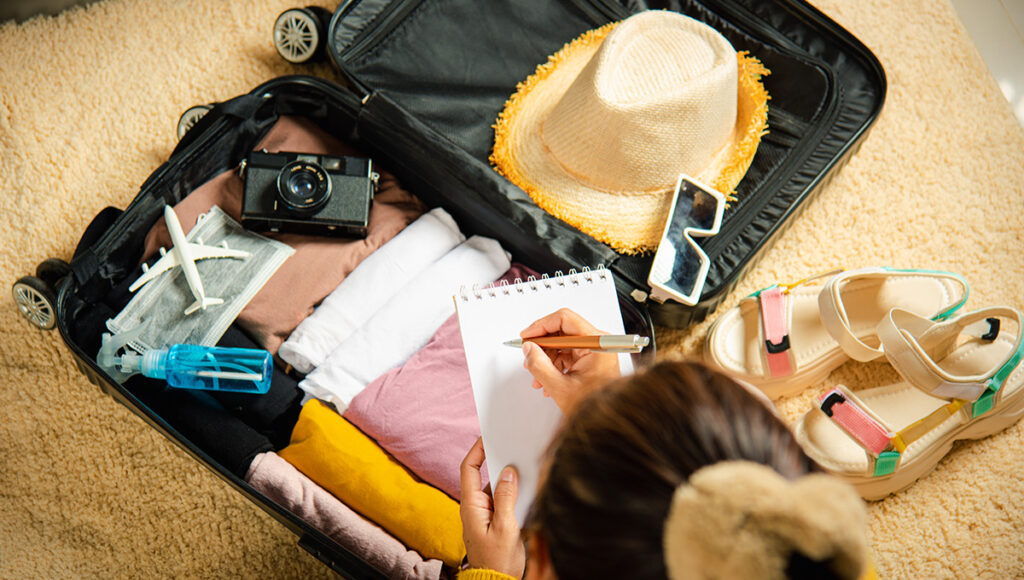 A person, seen from behind, is making a list in a small notebook while sitting next to an open suitcase filled with various travel essentials. Inside the black suitcase, neatly folded clothes in shades of pink, yellow, brown, and white are visible, along with a straw hat, a vintage camera, a small airplane model, a blue spray bottle, and sunglasses. To the right of the suitcase, a pair of light-colored, strapped sandals rests on the beige carpet. This image perfectly illustrates the process of preparing for a trip and figuring out what to pack to the Philippines, ensuring you have all your essentials.