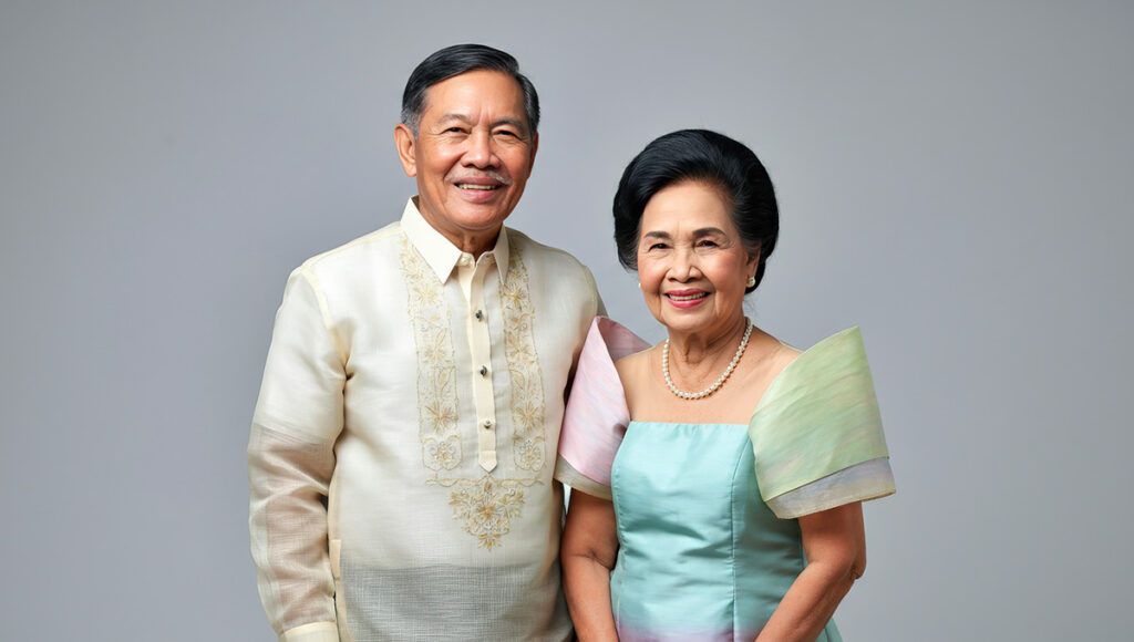 A Filipino man and woman stand side-by-side dressed in elegant Filipino traditional clothing, the man wearing a sheer embroidered Barong Tagalog, and the woman wearing a graceful Maria Clara gown, against a plain backdrop.