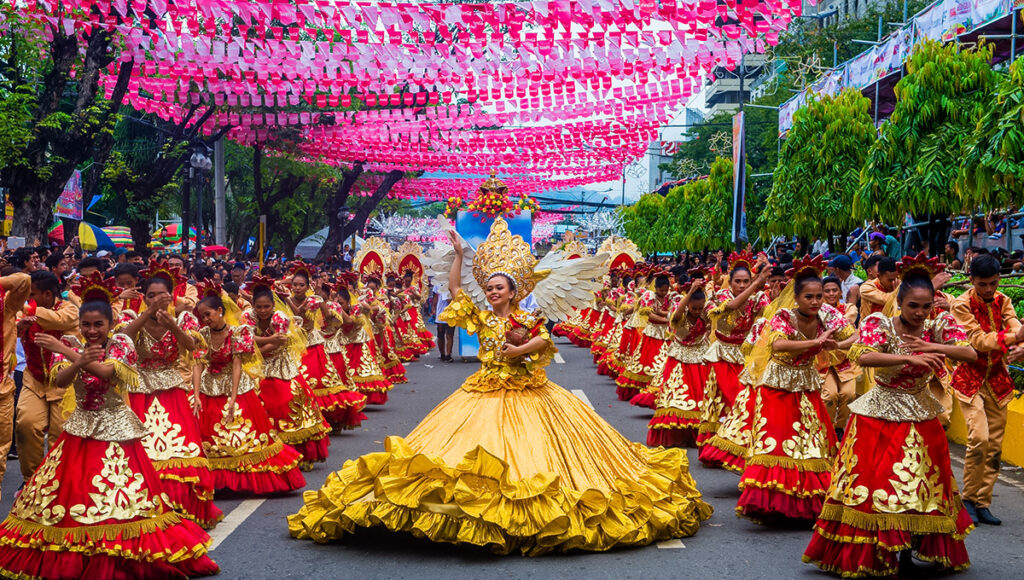 Cebu City Sinulog Festival Grand Parade featuring a lead dancer in a voluminous yellow gown and wings, surrounded by performers in red and gold costumes, with vibrant pink decorations strung across the street.