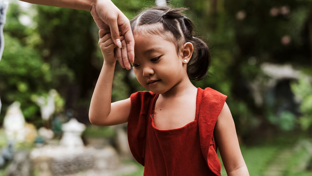 A close-up image of a young Filipino girl wearing a red top, looking down respectfully as an adult's hand gently rests on her forehead in the traditional "mano po" gesture, set against a blurred green, leafy outdoor background, showcasing Filipino culture.