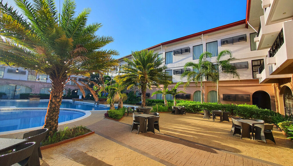 An outdoor resort area with a swimming pool, several palm trees, and dining tables and chairs on a sunny patio, suggesting the amenities of all inclusive resorts Philippines.