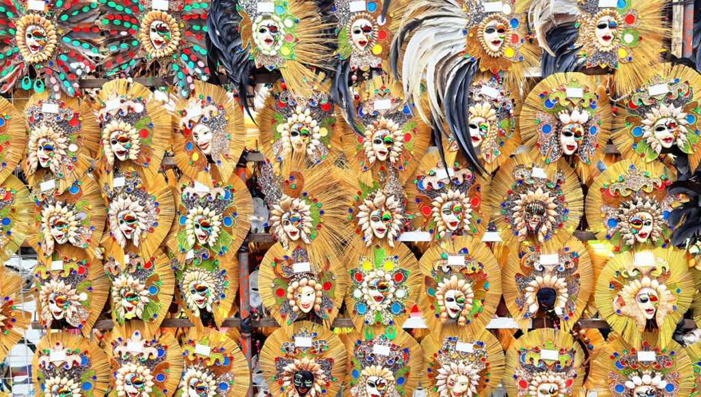 A dense, vibrant display of intricate, smiling masks from the MassKara Festival in Bacolod City, Philippines, featuring colorful feathers, beads, sequins, and other embellishments on golden, leaf-like backgrounds.