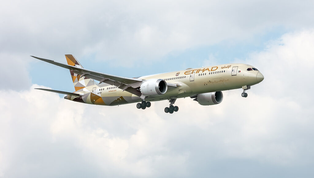 An Etihad Airways Boeing 787-9 Dreamliner, featuring its signature gold and brown 'Facets of Abu Dhabi' livery, is captured in mid-flight against a cloudy sky. The presence of this Middle Eastern carrier makes it relevant for those researching what airlines fly to the philippines from international destinations.