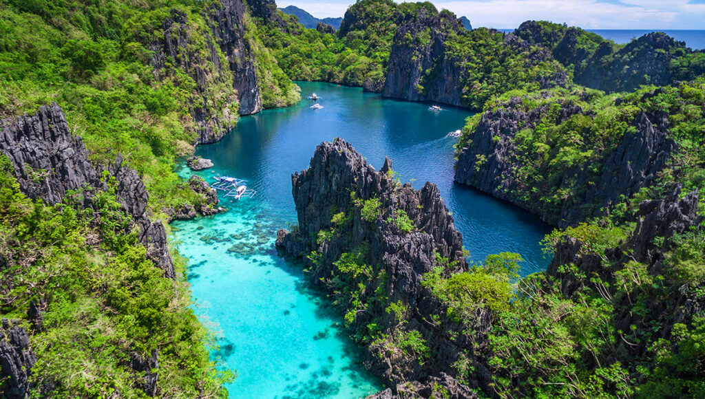 An aerial view reveals the stunning Big Lagoon in El Nido, Palawan, Philippines. Towering, jagged limestone karsts covered in lush green foliage frame a winding waterway that transitions from vibrant turquoise in the shallower foreground, where small boats are anchored, to a deeper indigo blue further back into the lagoon. A few small boats can be seen navigating the calm, clear waters. The scene is breathtaking, highlighting the natural beauty of the region.