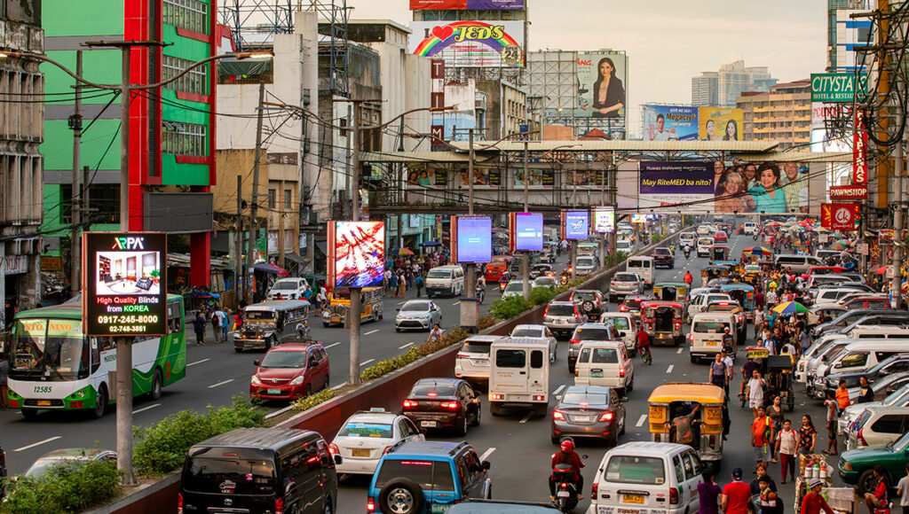 Crowded and busy highway street scene in Metro Manila featuring heavy traffic with cars, jeepneys, buses, and motorbikes, flanked by commercial buildings and large billboards. This image illustrates the dense, bustling nature of the city's main thoroughfares, providing context for travelers researching, "Is Manila safe?" regarding navigating transportation and urban density.