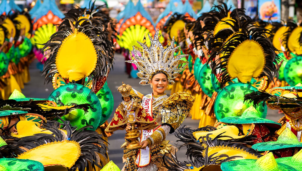 Image of Cebu City Sinulog Festival Queen holding the Santo Niño icon, surrounded by performers in elaborate yellow, black, and bright green costumes during the Sinulog street parade.