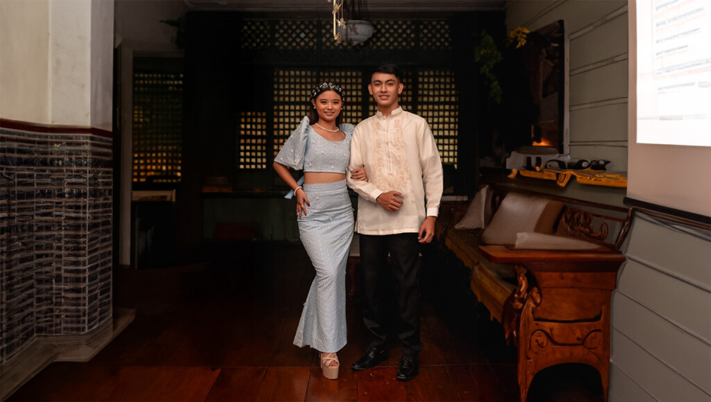 A young Filipino couple stands together indoors; the woman wears a light blue modern Filipiniana two-piece outfit with a mermaid skirt and puff sleeves, and the man wears a traditional Barong Tagalog shirt. They are posing in a dimly lit, elegant room with dark wooden floors and antique furniture, highlighting Filipino culture.