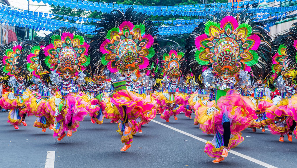 A group of performers dancing in the street during the MassKara Festival in Bacolod City, wearing elaborate, towering headdresses adorned with bright pink, green, and black feathers, coupled with voluminous pink and yellow ruffled skirts and matching smiling masks.