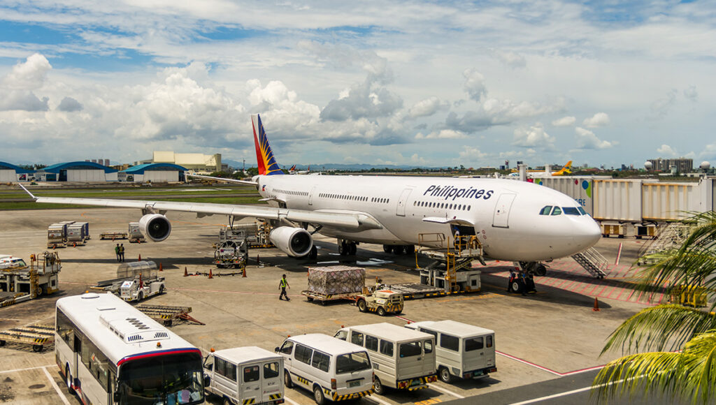 A Philippine Airlines aircraft, identified by "Philippines" on its fuselage and the airline's logo on the tail, is parked at a busy airport gate under a partly cloudy sky. Ground crew and various vehicles, including luggage carts, buses, and vans, are actively working around the plane, suggesting preparations for departure or arrival. The scene makes one wonder about international connections, prompting the question: When will Philippine Airlines resume flights to London?