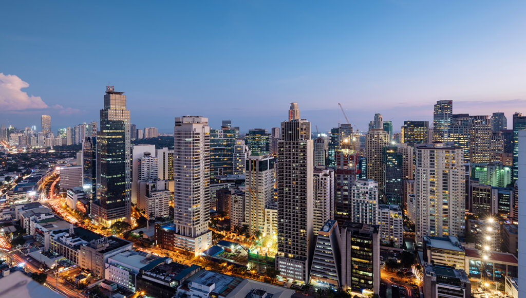 An expansive aerial view captures the vibrant Makati skyline in Manila, Philippines, at dusk. Numerous illuminated skyscrapers of varying heights dominate the frame, their windows glowing with warm light against the deep blue twilight sky. Below, busy streets are marked by streaks of red and white from car lights, indicating the bustling urban environment. The city lights stretch into the distance, showcasing the dense development of this major financial district.