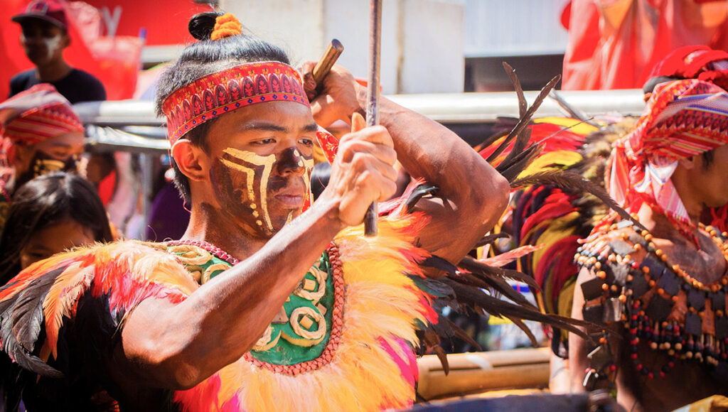 A male performer in face paint and a feathered costume beats a drum with a stick during the Dinagyang Festival celebration.