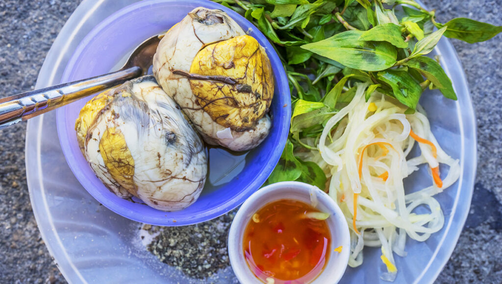 Close-up, overhead view of Balut, the Filipino fertilized duck egg, served in a purple bowl alongside fresh mint leaves, pickled papaya strips, ground pepper, and chili dipping sauce. This controversial delicacy is one of the most talked-about fun facts about the Philippines' unique and adventurous cuisine.