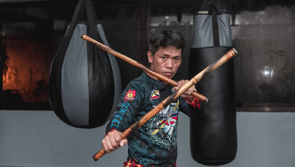 A focused portrait of a Filipino man wearing a dark martial arts uniform with various patches, holding two wooden rattan sticks crossed in front of him, demonstrating the Arnis martial art, an important part of Filipino culture. He stands in a training space with large black punching bags visible in the blurred background.