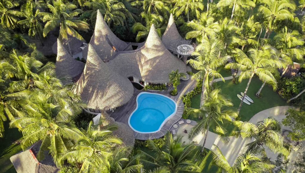 A high-angle aerial view of the Nay Palad Hideaway, a luxury eco-resort on Siargao Island, Philippines. The resort features distinctive, high-peaked thatched-roof structures grouped around a vibrant blue swimming pool and surrounded by lush green tropical foliage and palm trees.