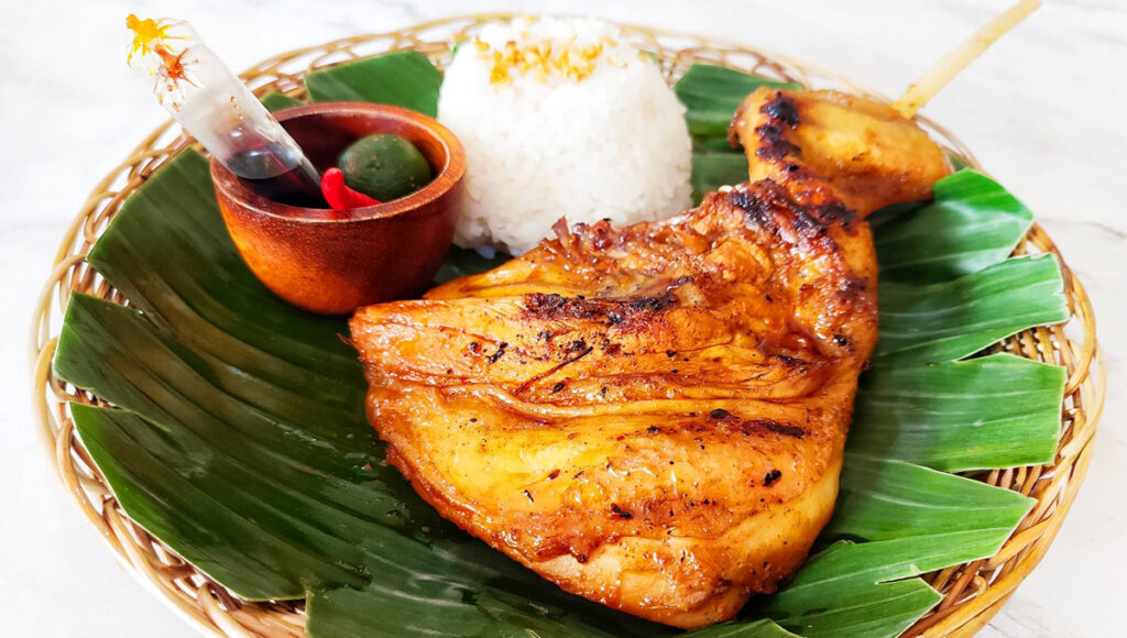 A serving of Ilonggo Chicken Inasal, consisting of a large, grilled chicken quarter with crispy skin, served on a banana leaf in a woven plate with a scoop of garlic rice, and a small wooden bowl of soy sauce, chili, and calamansi for dipping.