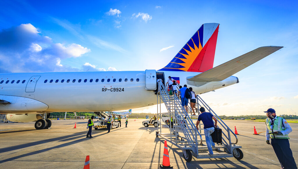 Passengers are boarding a Philippine Airlines aircraft via mobile stairs on a sunny day with a bright blue sky and some clouds. The airline's distinctive sun logo is visible on the tail fin. Ground crew members are also present near the aircraft, and orange cones are set up on the tarmac.