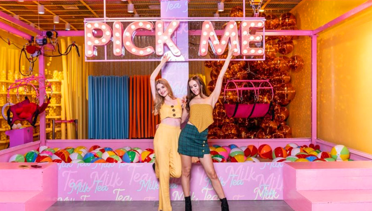 Two young women posing playfully in a vibrant, pink and yellow interactive museum exhibit with a large, illuminated "PICK ME" sign and colourful balls, suggesting that the fun attractions prove is Manila worth visiting.
