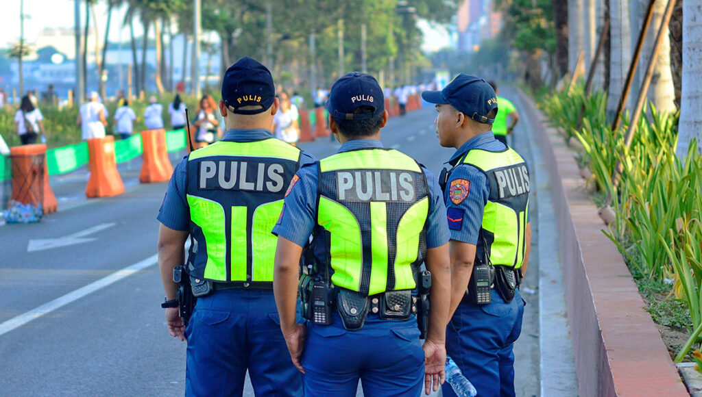 Three uniformed members of the Philippine National Police (PULIS) wearing high-visibility vests stand guard on a wide public street in Manila, with a crowd of people and temporary barricades visible in the background. This image demonstrates the high level of visible security presence, directly answering the question, "Is Manila safe?"