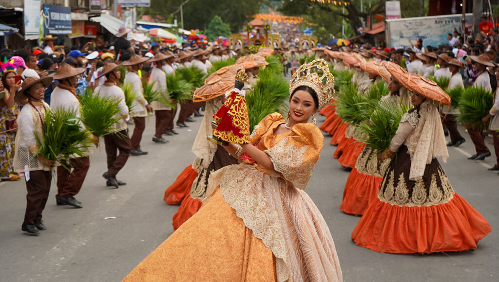 Cebu City Sinulog Festival street parade featuring a Festival Queen in a beige and orange Filipiniana gown, holding the Santo Niño icon, surrounded by dancers wearing brown hats and traditional costumes holding palm fronds.