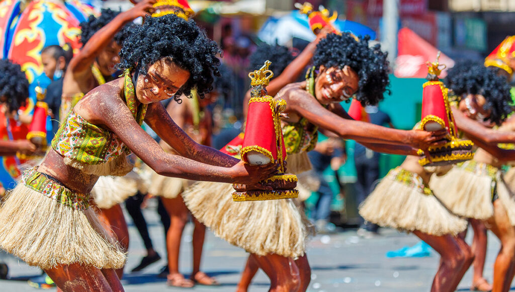 Dark-skinned performers in straw skirts, short tops, and black curly wigs dance in the street while holding small Santo Niño replicas during the Dinagyang Festival.