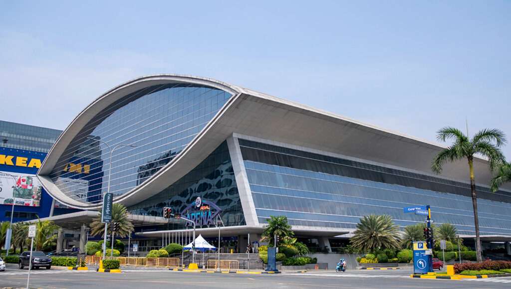Exterior view of the enormous SM Mall of Asia (SM MOA) complex in Pasay City, Philippines, featuring its modern, curved glass facade and surrounding palm trees on a sunny day. The mall's massive size is one of the incredible fun facts about the Philippines' unique mall culture and its role as a social hub.