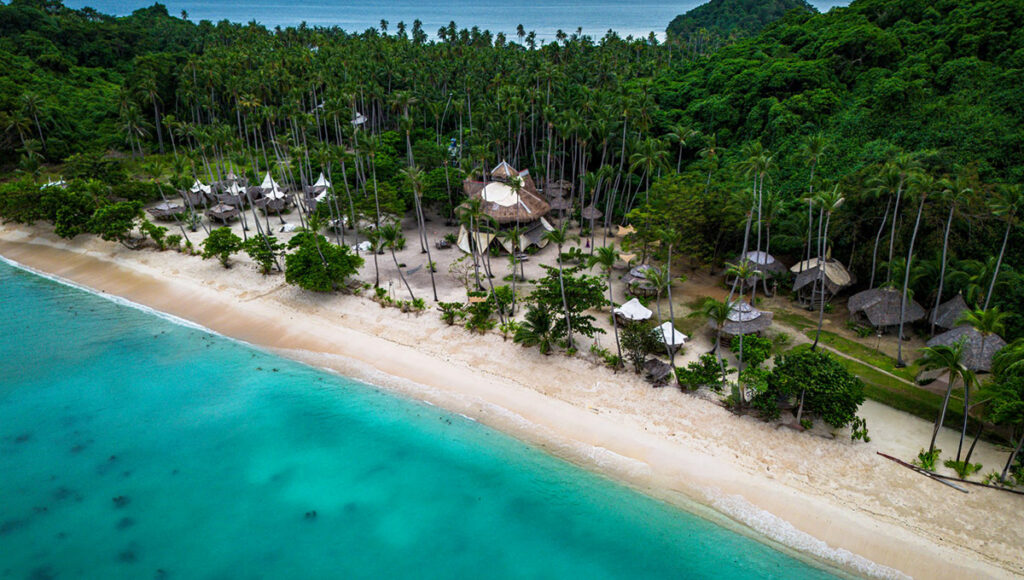 An aerial view of a beautiful, secluded cove in El Nido, Palawan, Philippines, featuring a long stretch of white-sand beach and vibrant turquoise water. The beach is backed by dense tropical jungle and palm trees, where the thatched-roof accommodations of a luxury resort, specifically an El Nido Resorts property, are nestled.