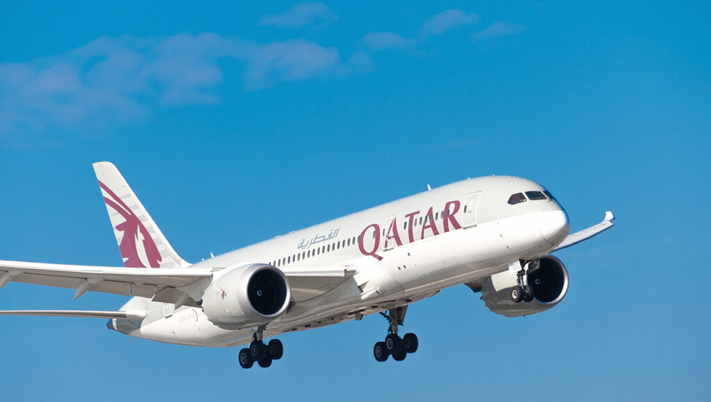 A large Qatar Airways Boeing 787 Dreamliner airplane, painted white with a distinctive purple and maroon tail logo, is captured ascending against a bright blue sky with a few wispy white clouds. As a major international carrier, this aircraft is representative of the types of flights that answer the question: what airlines fly to the Philippines?