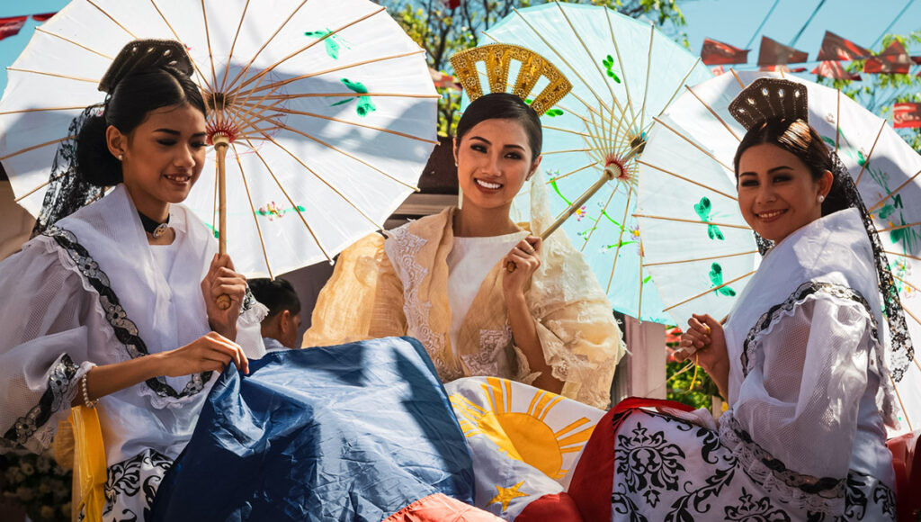 Three Filipina women in traditional white and black Baro't Saya attire, holding white paper parasols, sitting on a float adorned with the Philippine flag during the Cebu City Sinulog Festival parade.