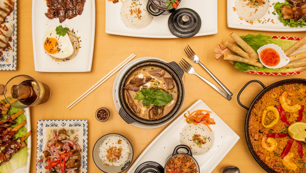 A high-angle, overhead shot of a table completely covered with a variety of colorful and appetizing dishes, including beef kare-kare (peanut stew), rice, lumpia (spring rolls), meat skewers, and seafood paella, showcasing the rich variety of Filipino food and Filipino culture.