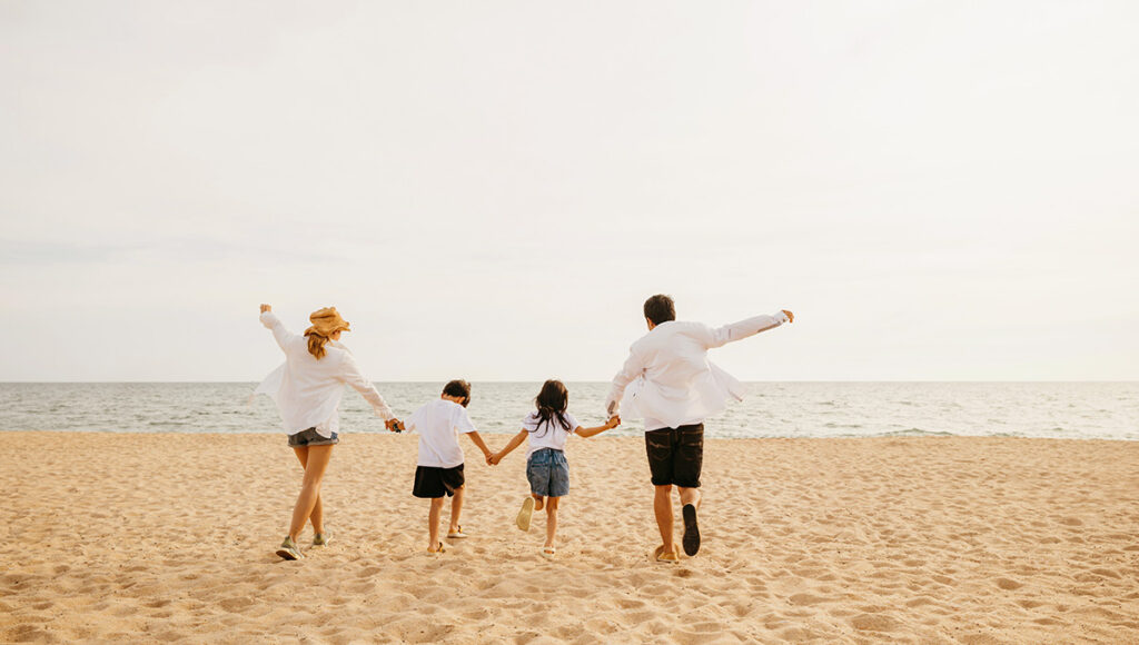 A family of four, including two adults and two children, holds hands and runs toward the ocean on a sandy beach, symbolising a joyful vacation experience at all-inclusive resorts Philippines.