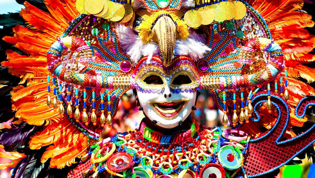 A close-up of a performer wearing an incredibly vibrant and ornate costume, featuring a white smiling mask with gold trim around the eyes and red lips, surrounded by a large headdress with bright orange, blue, green, and pink feathers, dangling beads, and shiny gold accents, during the MassKara Festival in Bacolod City.
