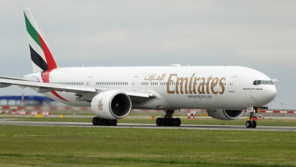 A side-on view of a white Emirates Boeing 777-300ER airplane with the distinctive red, green, and black tail livery, taxiing on a runway. The presence of this major international carrier, which flies long-haul routes, is relevant for those searching for what airlines fly to the Philippines.