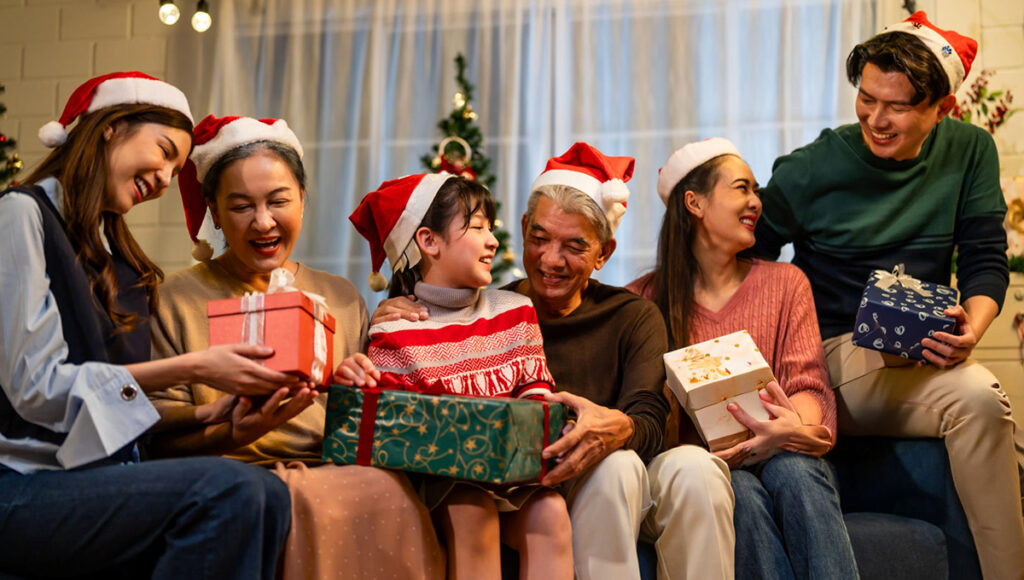 A Filipino family wearing Santa hats is gathered in a living room decorated for Christmas, exchanging wrapped presents near a large tree, showcasing Filipino Christmas traditions.