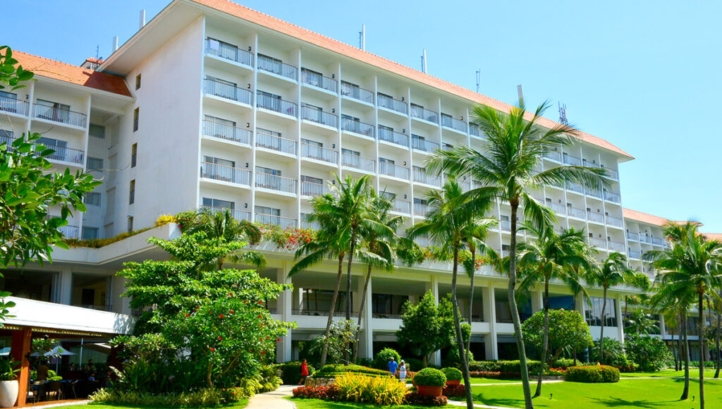 A low-angle exterior view of the Shangri-La Mactan, Cebu, a large, multi-story white hotel building with an orange-tiled roof and multiple balconies. The foreground features a vibrant, well-manicured tropical garden with lush green lawn, flower beds, and tall palm trees under a bright blue sky.