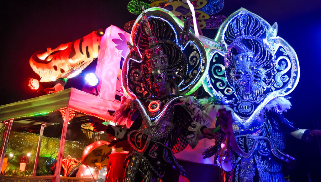 Two parade performers in highly elaborate, futuristic costumes with large, glowing silver and black headdresses illuminated by bright blue, red, and pink neon lights, representing the electric masskara street dance during the nighttime MassKara Festival in Bacolod City.