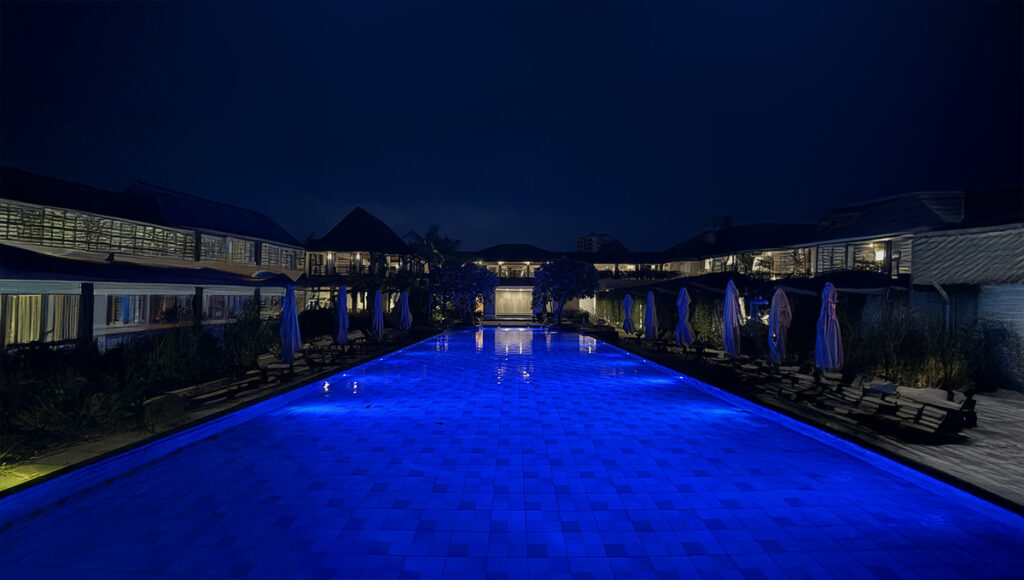 A wide, panoramic night view of the main pool area at the Crimson Resort and Spa Mactan, Cebu. The long, rectangular swimming pool glows with an intense blue light, surrounded by resort buildings on both sides with warm interior lighting and rows of sun loungers covered by folded umbrellas.