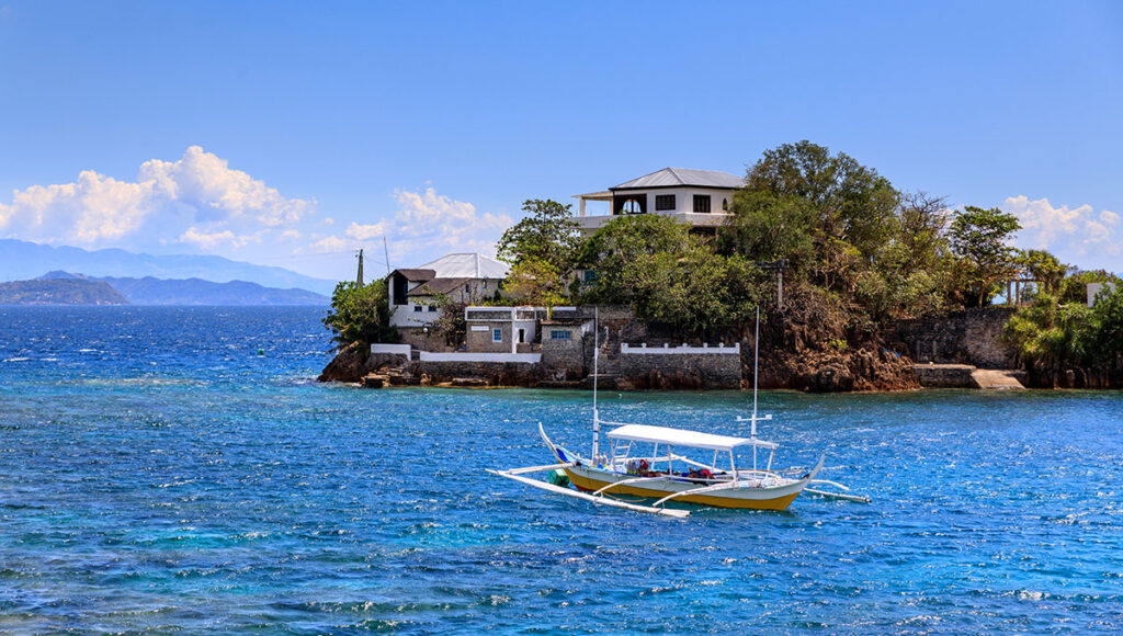 A small outrigger boat (banca) floats near a rocky shoreline with houses on a hill in Anilao, Batangas.