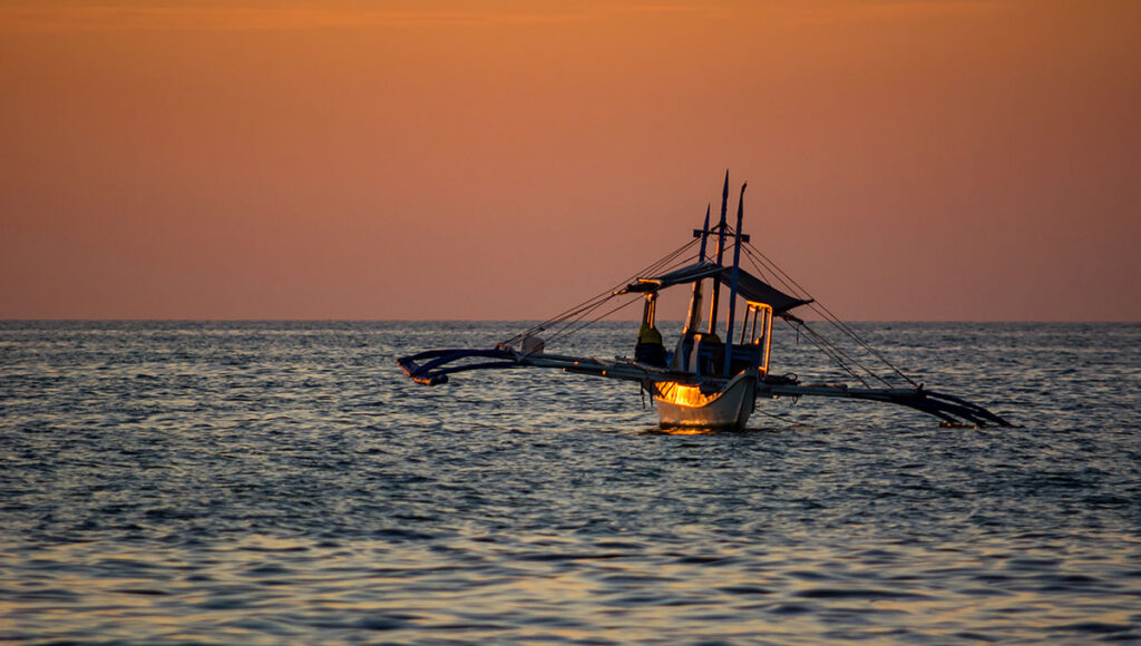A silhouette of a traditional Bangka boat (outrigger canoe) floating on the calm sea during an orange and pink sunset, symbolizing the island beauty and maritime culture that is what is Philippines famous for.
