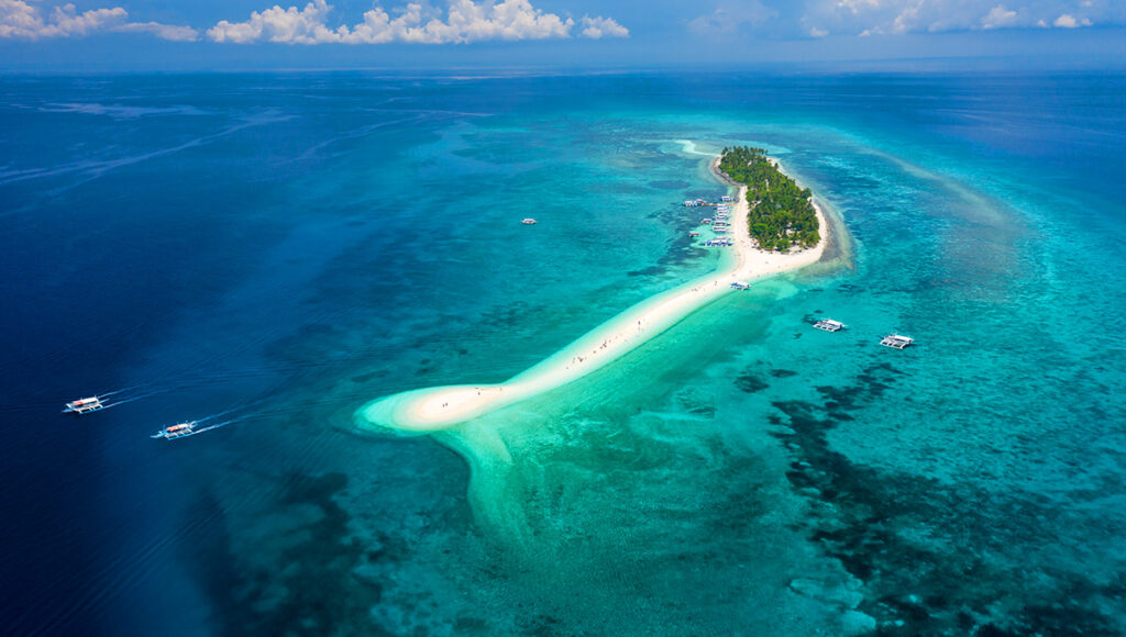 Aerial view of a stunning white sandbar and small palm island, surrounded by turquoise water, often found during island hopping Philippines tours in the Cebu region.