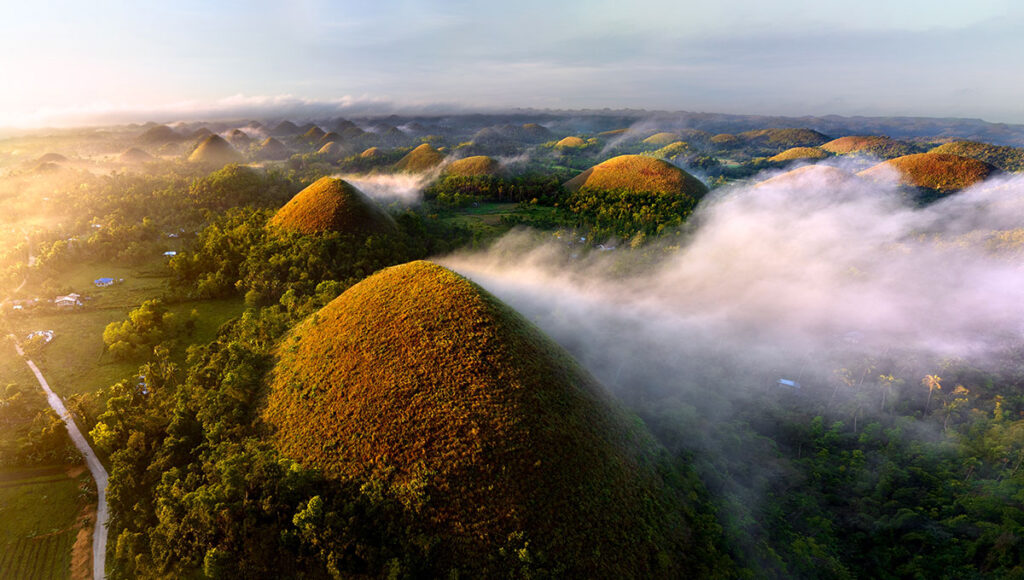 Aerial view of the Chocolate Hills in Bohol, Philippines, covered in green grass and shrouded in mist during sunrise, highlighting another natural wonder of what is Philippines famous for.