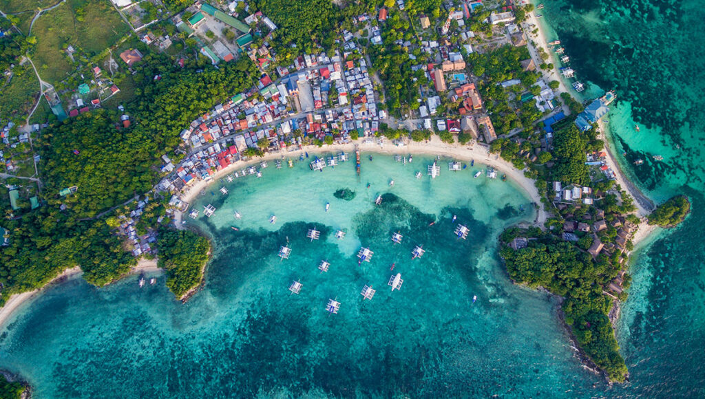 Overhead aerial view of the densely built-up beach and bay of Malapascua Island, filled with boats and surrounded by turquoise water.