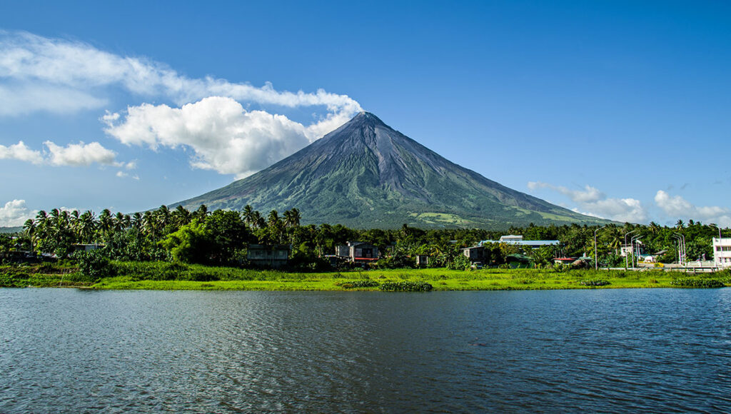 The perfectly conical and majestic Mayon Volcano in Albay, surrounded by lush greenery and a foreground of water, representing the stunning natural geography and volcanoes that is what is Philippines famous for.