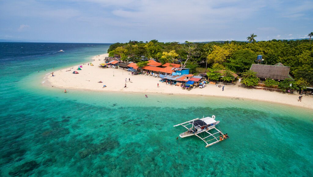 A sandy beach lined with local buildings and a boat in the clear turquoise water of Moalboal, Cebu, a popular Philippines scuba diving spot.