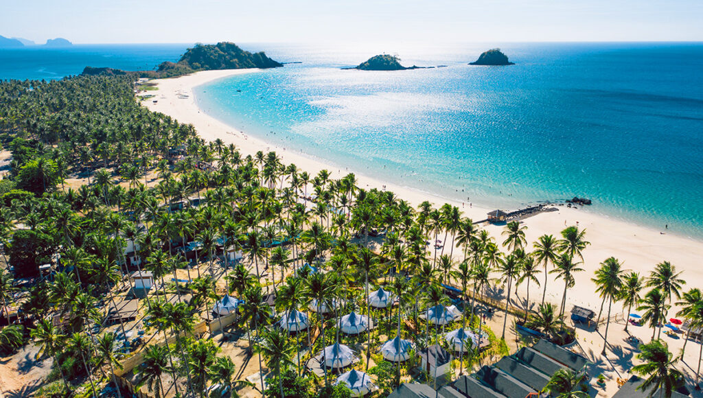 Vibrant aerial view of a Palawan beach with a long stretch of white sand, lush palm trees, and crystal-clear turquoise water. An iconic Philippines island destination perfect for Philippines travel.