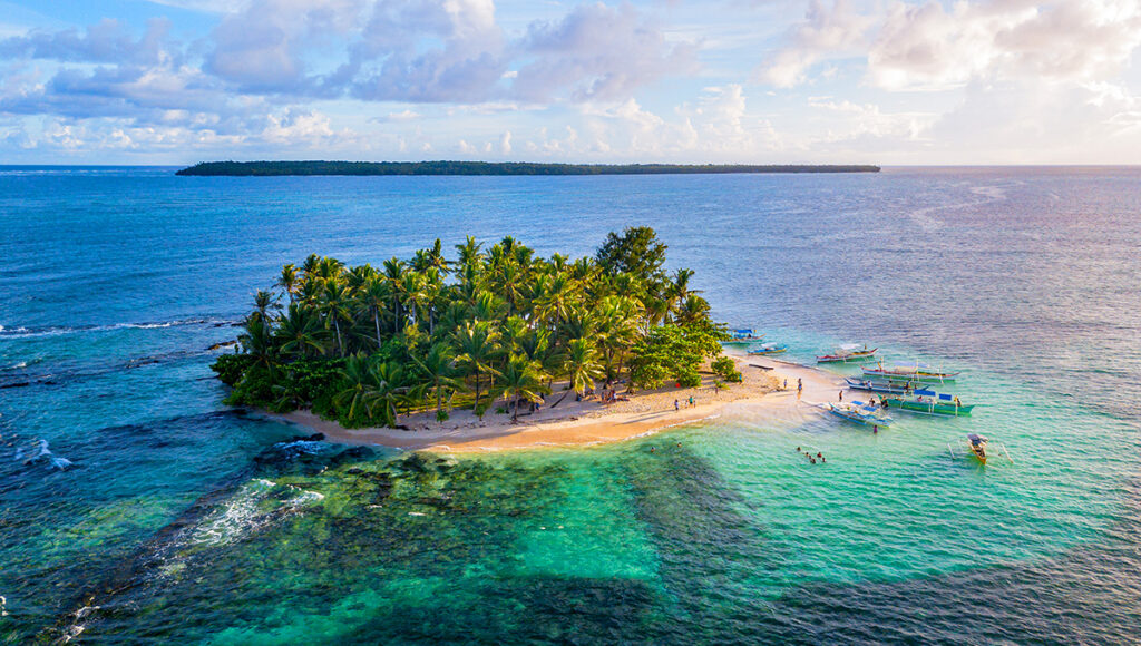 Aerial view of Guyam Island off the coast of Siargao, a tiny tropical oasis covered in palm trees with boats anchored in the surrounding shallow, turquoise waters. A classic Philippines island-hopping scene.